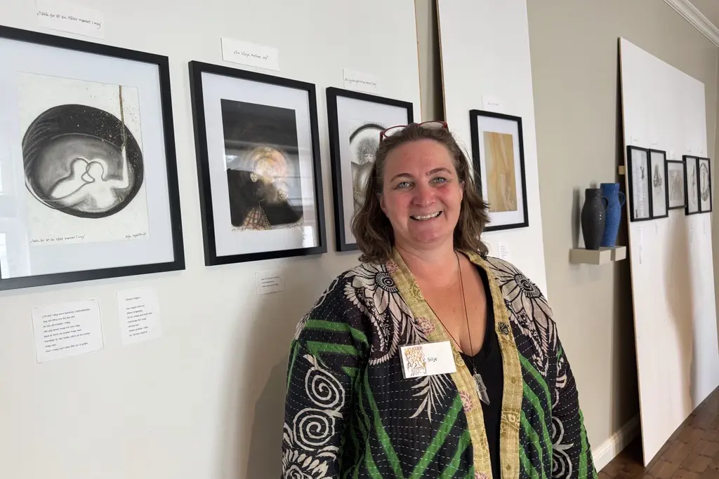 Silje Engbråten står innendørs foran en vegg med sine innrammede kunstverk. Flere bilder henger på rekke langs veggen, og små tekstlapper er plassert ved siden av verkene.