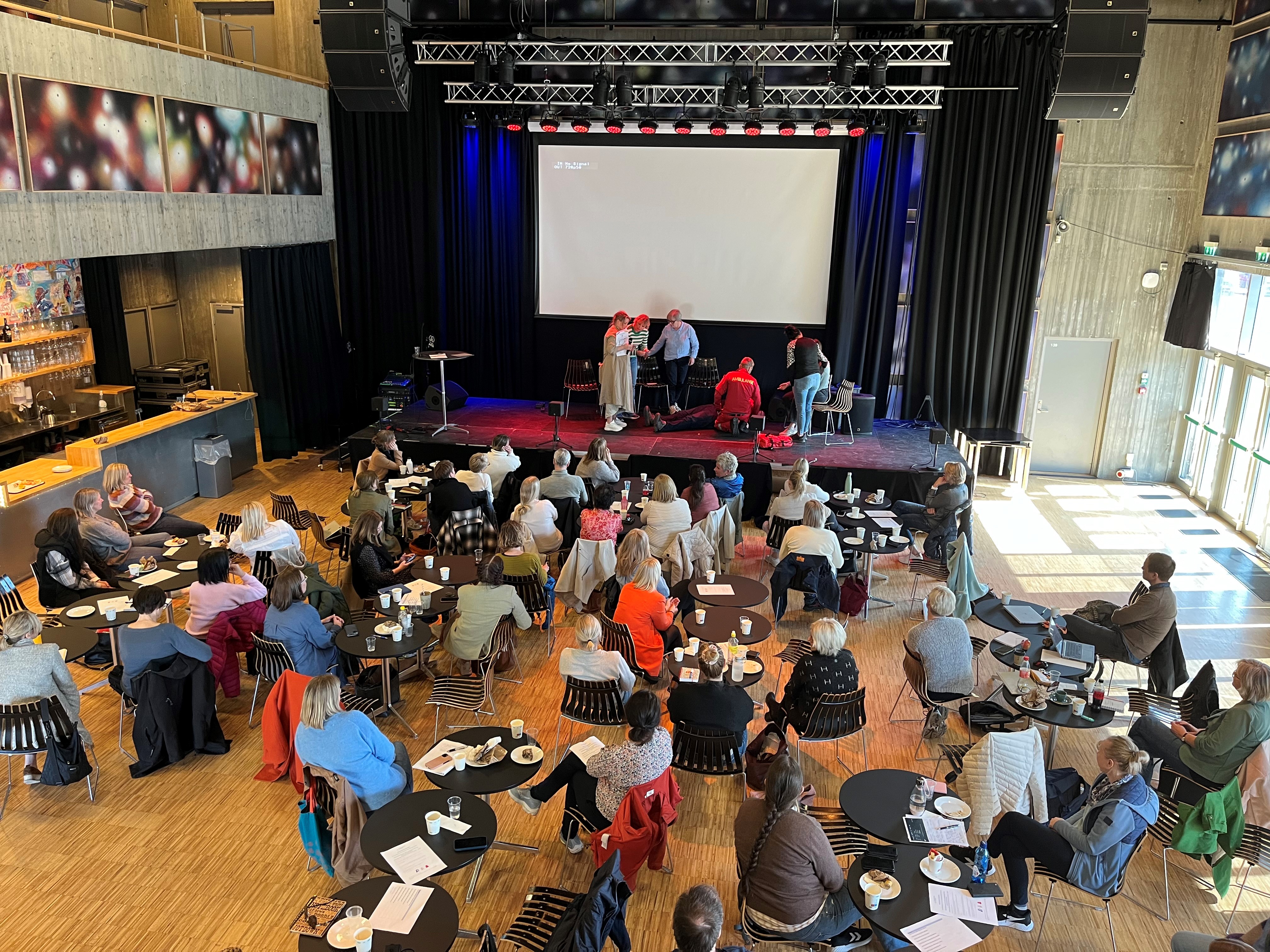 En gruppe mennesker som sitter i et rom med en scene og en skjerm