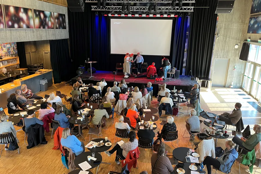 En gruppe mennesker som sitter i et rom med en scene og en skjerm